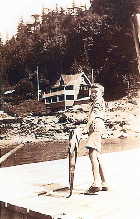 Tommy Huggan standing with a fish on the government dock in Mt. Gardner. The tea room is visible in the background. ca. 1940’s