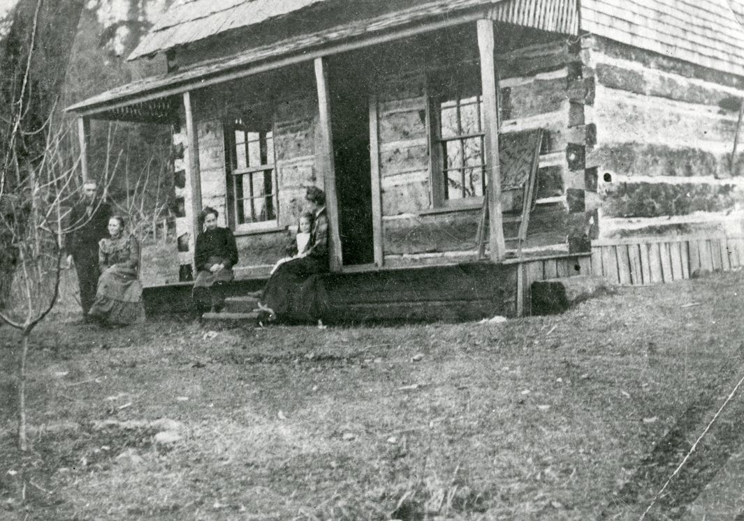 Photo of the Galbraith family cabin circa 1906, a log structure with a pitched roof and visible chinking between horizontal logs. Two windows flank a central door. Three women in dresses sit on the porch edge, one beside a child. A man stands to the left near a support beam. The cabin sits in a grassy, dry area with sparse trees in the background, some without foliage.