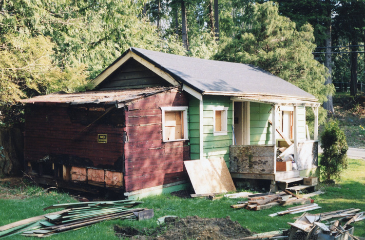 Pre-restoration view of the Higgins Loggers Cabin—a small, single-story structure in a grassy, wooded area. The main cabin is green with intact siding, a porch, and wooden steps. A worn red addition on the left shows missing panels and exposed insulation. Construction debris is scattered in front, and a road runs behind the cabin.