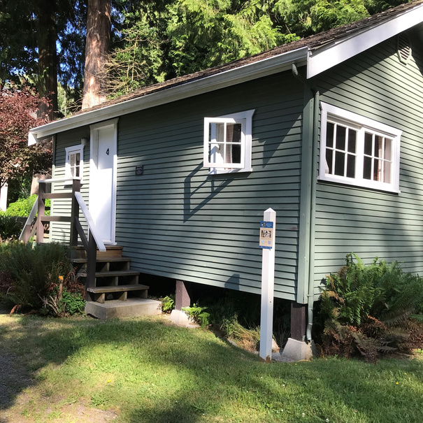 Restored Higgins Cabin as of 2000. A small, green-painted, single-story cabin with horizontal wood paneling, elevated on concrete blocks. It features two front windows, a white door marked with the number '4,' and wooden steps with a handrail. The cabin sits in a grassy area with ferns, surrounded by tall trees. A white post with a Bowen Island Heritage Trail sign stands nearby.