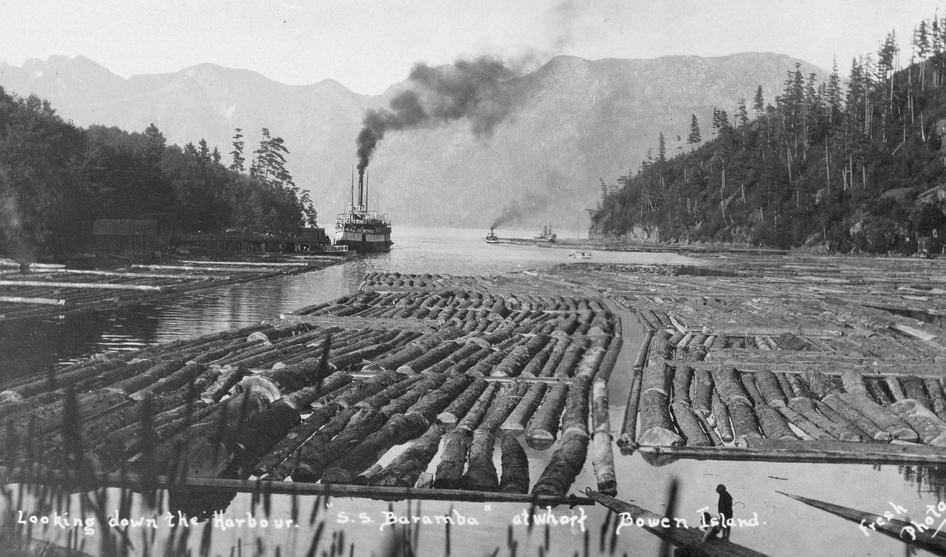 Harbour scene at Snug cove, Bowen Island with multiple log rafts floating in the water. The S.S. Baramba is docked at a wharf on the far left. A person stands near the water’s edge observing the logs. Forested mountains rise on both sides of the harbour, and a smaller vessel is visible in the distance.