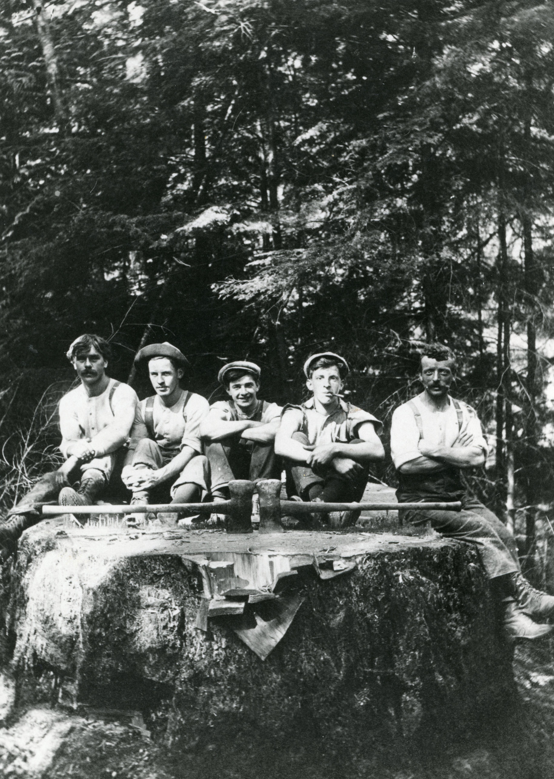 Black and white photo of five men seated on a large tree stump in the forest at Grafton Bay. Two axes lie across the stump with blades facing each other. The men, dressed in work clothes with suspenders, caps, and boots, appear to be resting. Identified left to right: Hans Lehr, Cameron Smith, Bobby Greenwell, George Dorman, and Fred Billington.