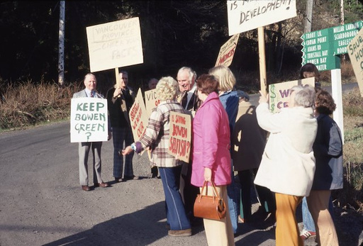 Photo from 1977 showing a group of about ten people standing on the roadside protesting the Stan James development. They hold signs, including one reading 'KEEP BOWEN GREEN.' Dressed in casual clothes and jackets, the group stands close together. The road is bordered by grass and vegetation, with a signpost and multiple directional signs visible in the background.