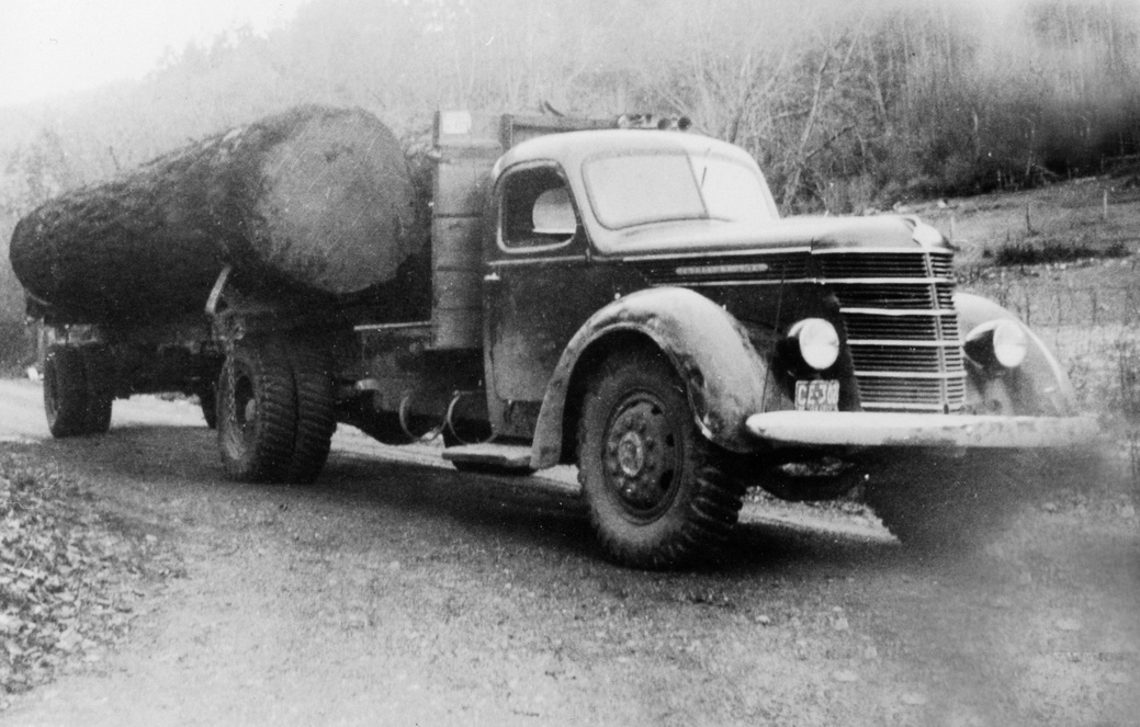 Logging truck near Killarney Lake, dated between 1930 and 1950, transporting large logs on a dirt road. The truck faces right, featuring a prominent front grill with horizontal slats, round headlights, large treaded tires, and a flatbed. Trees and a gentle slope form the background.