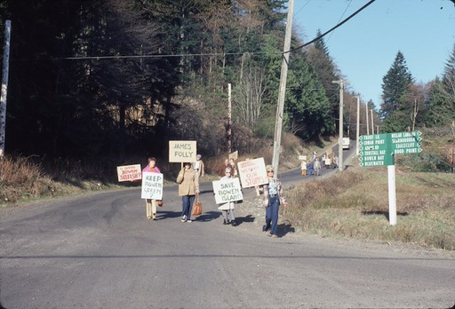 Photo from 1977 showing a group protesting the Stan James development while walking along a curved paved road lined with trees and utility poles. Four people in the foreground hold signs reading 'Keep Bowen Green,' 'James Folly,' and 'Save Bowen Island.' More protesters with signs follow in the background. A green directional signpost is visible on the right side of the road.