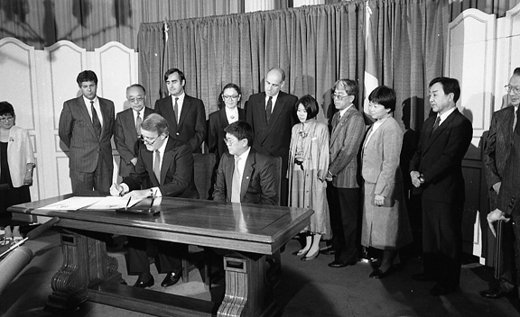 A group of Japanese dignitaries watches Canadian Prime Minister Brian Mulroney signing the document.
