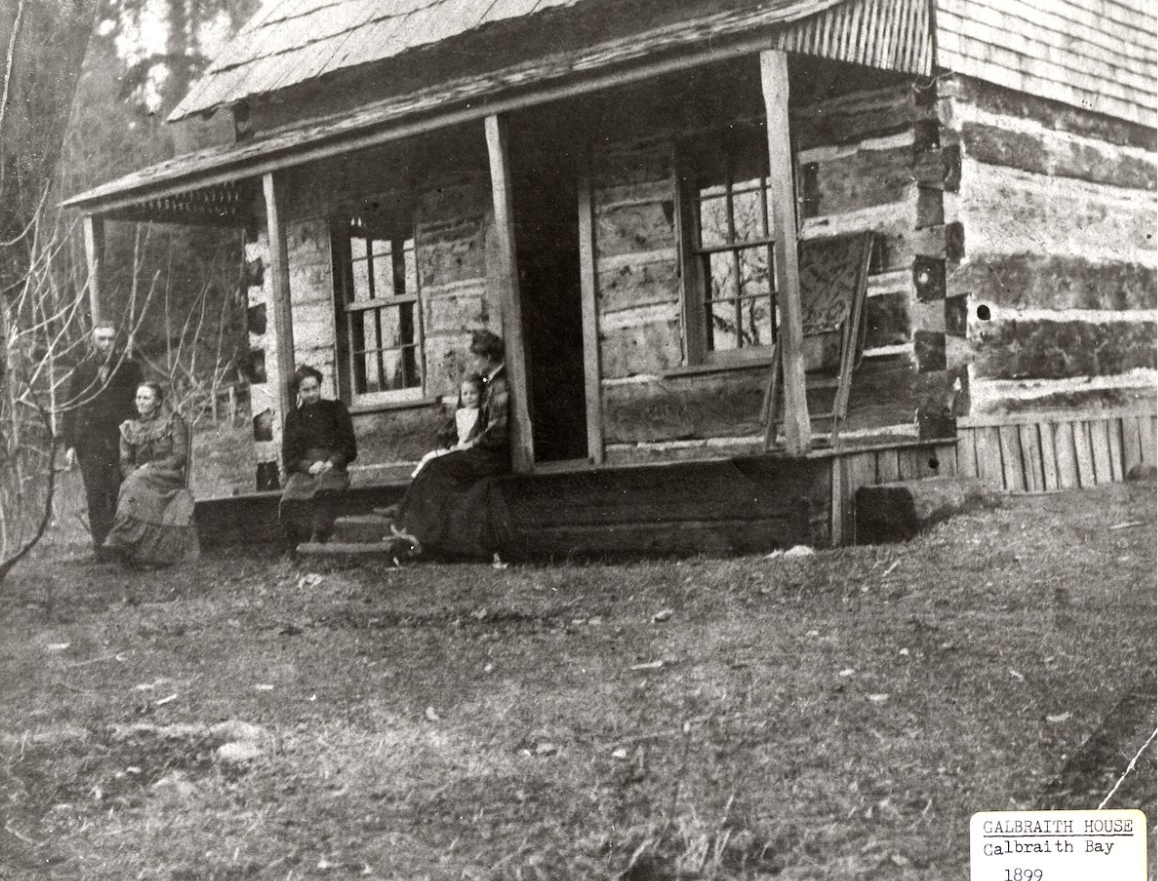 A man, three women and a child cluster on the porch of a rough-hewn log cabin.