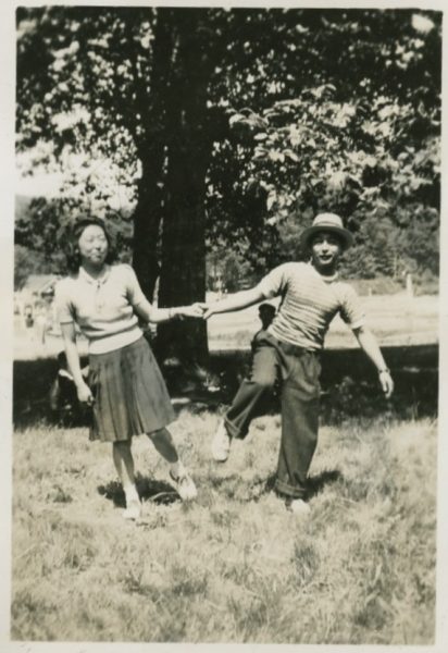 A young man and woman holding hands, leaning apart in a field, the the man's leg lifted playfully in the air.