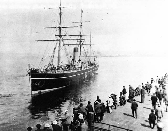 People line up along the edge of a pier and a wooden tall ship floats in, sails down. A smokestack indicates it is a steamer as well.
