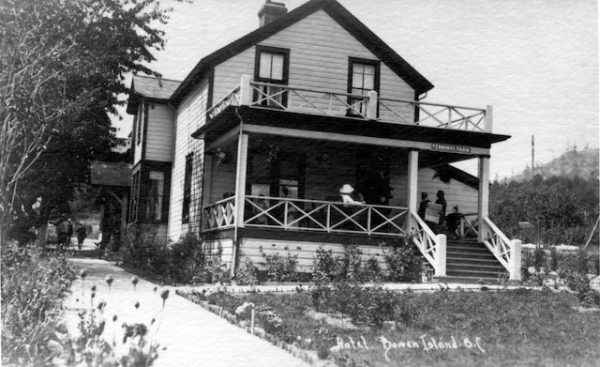 A larege two story wooden house with front and side entrances and with people seated on a large covered porch