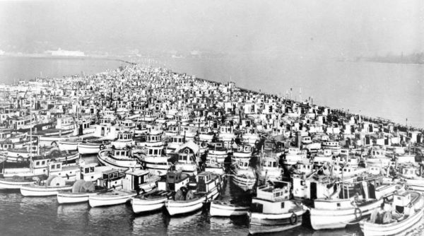 A great number of wooden boats in the water packed against a breakwater with no docks between them.
